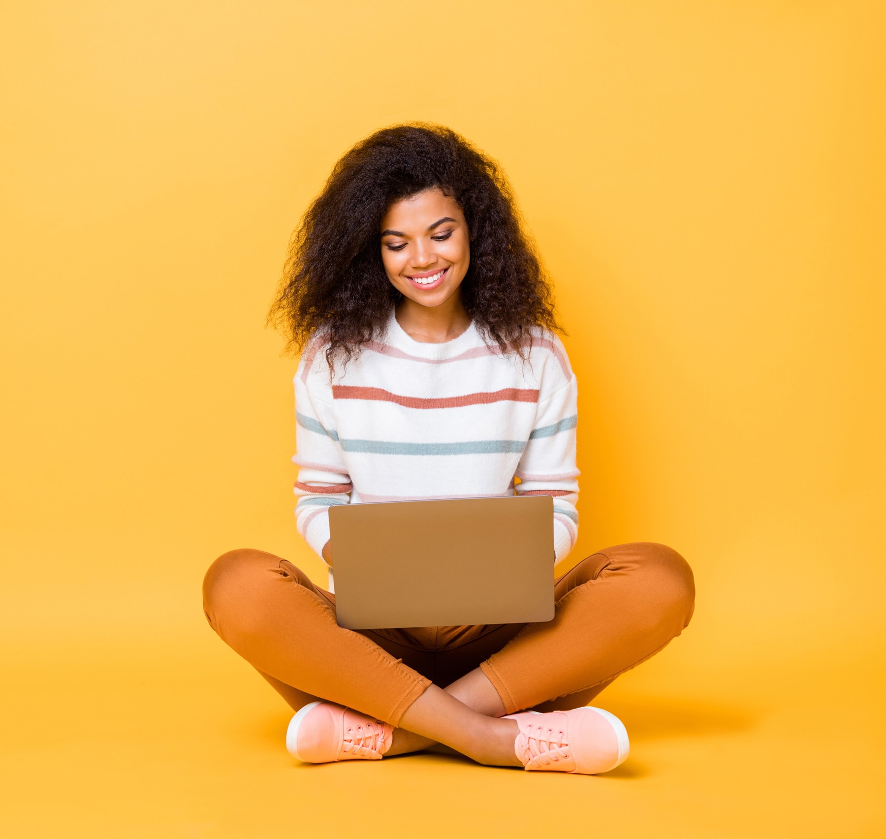 woman of color sits before an orange background smiling and working on her laptop