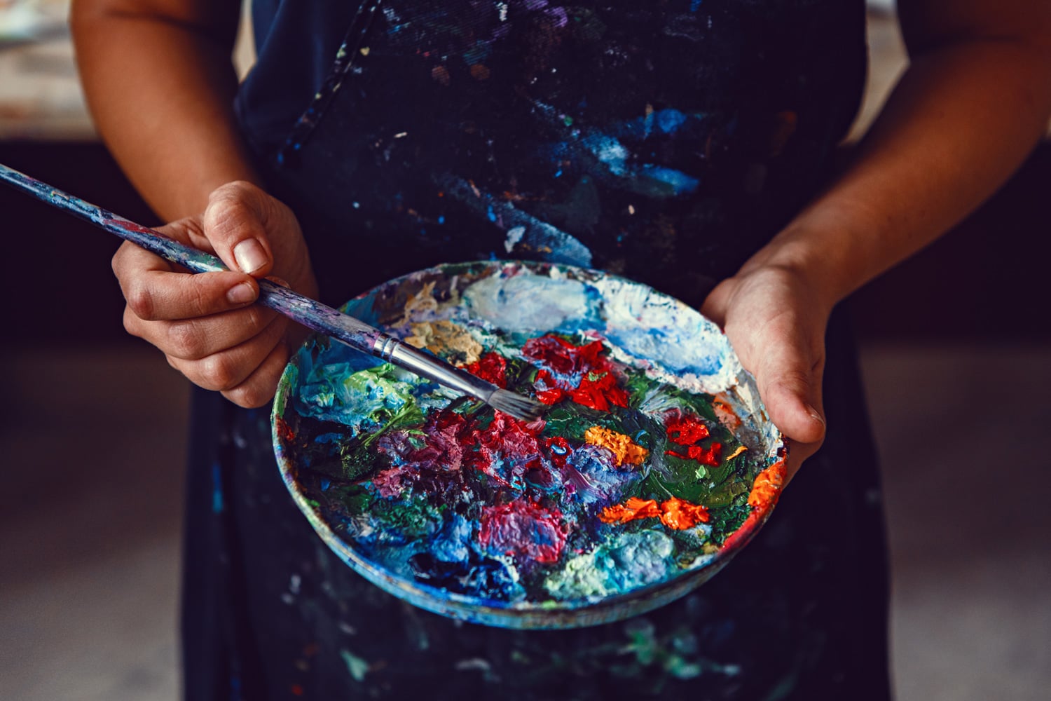 A painter holds their palette in both hands, and paint of all colors can be seen across the plate like a rainbow starry night. They are holding a long, skinny paint brush and wearing an apron.