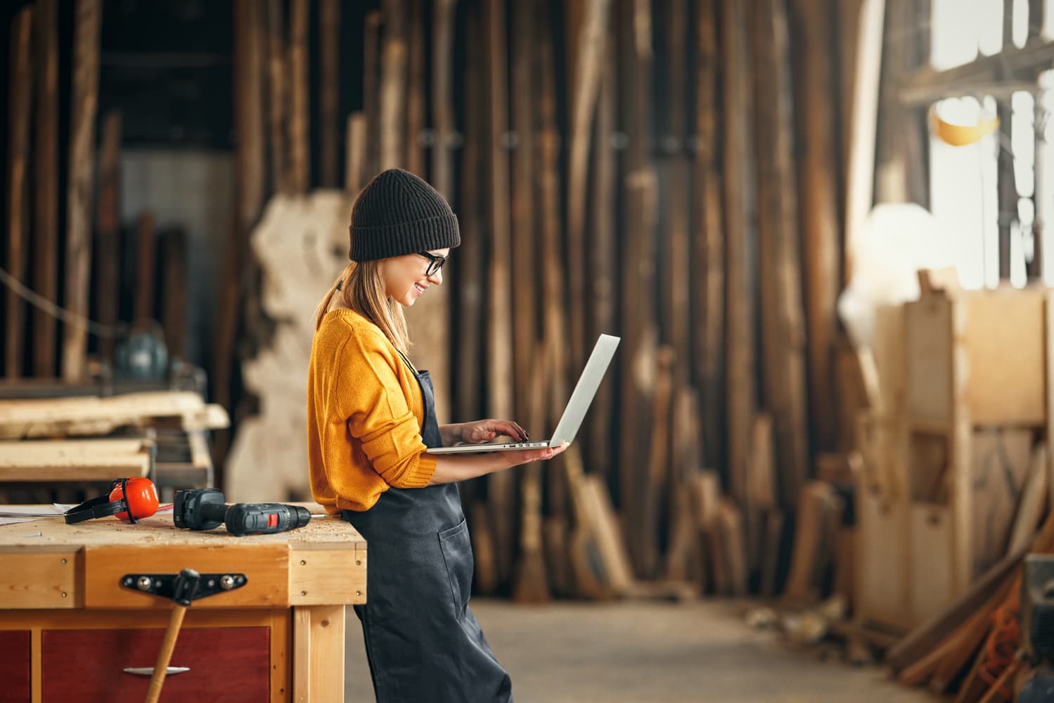 A person can be seen leaning against a work bench, holding their laptop up while working on it. There are tools around the workshop and the person is wearing a yellow shirt, beanie, and apron.