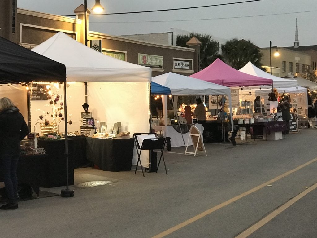 A row of canopies can be seen going down the street. It is evening and the canopies are multiple colors. All sorts of arts and crafts can be seen on the tables at this event on Race Street in Fort Worth