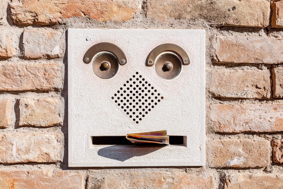 A metal mailbox is imbedded in a pale beige brick wall. The mailbox has hardware that looks like eyebrows, eyes, nose, and a mouth. There is a letter sticking halfway out of the mouth.