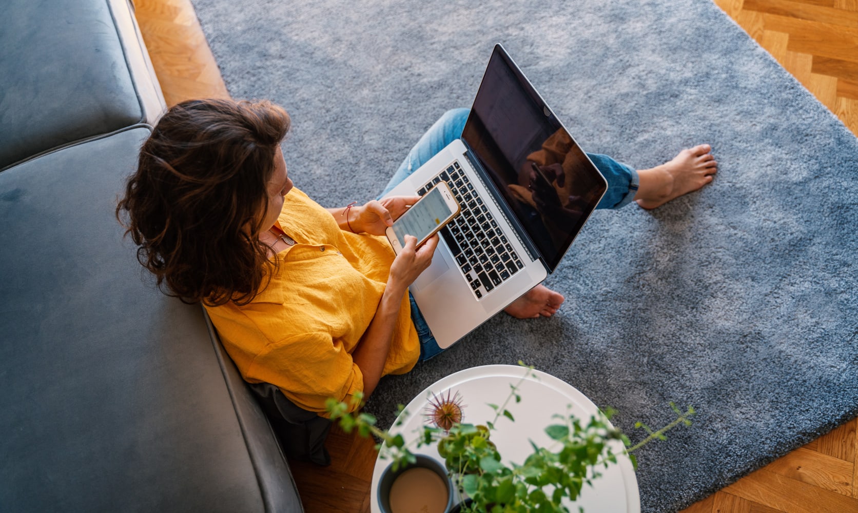 A person with dark, mid-length hair sits on the floor, leaning against a blue couch. They are checking their phone while working on their laptop, sitting on a plush blue rug against an orange toned wood floor.
