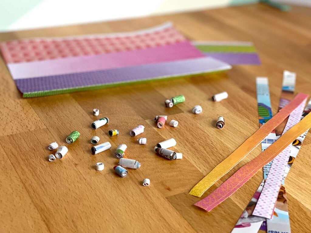 Decorative paper and cut paper strips sit around a pile of finished paper beads all from the Recycled Paper Beads Kit by 4M.