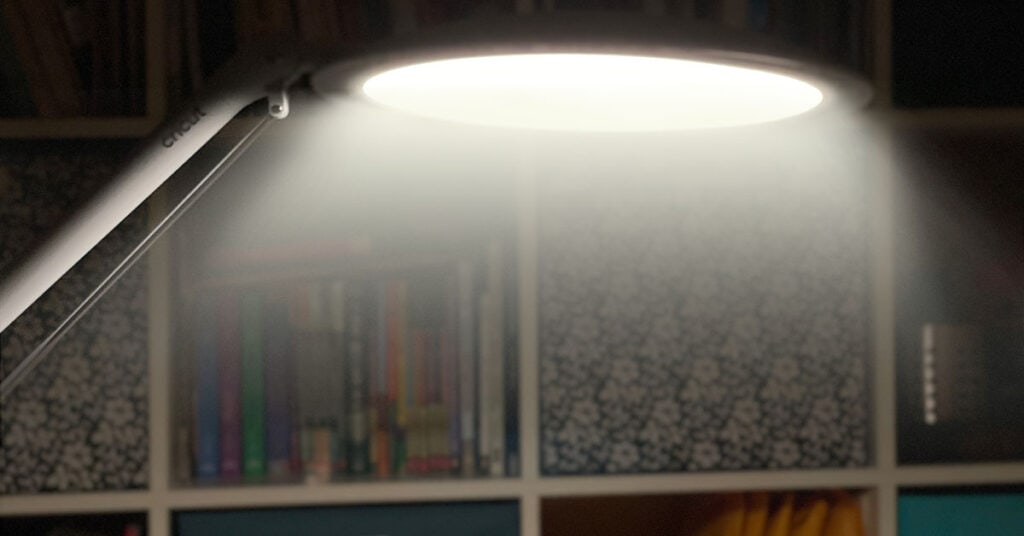 Cricut Bright 360 Lamp is on in an otherwise dark room with shelves of craft supplies in the background