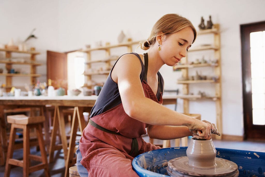 a person in a pink apron works on a vase at a potters wheel. Her hair is pulled back and there are shelves of ceramic art in the background.