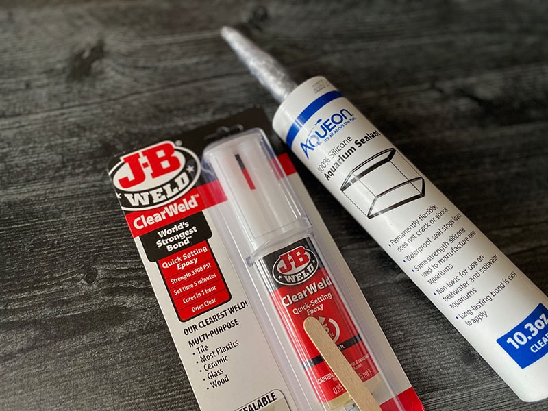 JB Clear Weld Syringe and Aquarium Safe Silicone resting on a dark wood background.