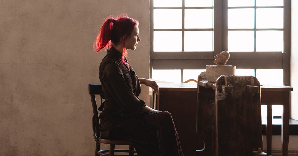 An artist sits at her desk partially illuminated by the window. She is mostly a silhouette swallowed up by the darkness.