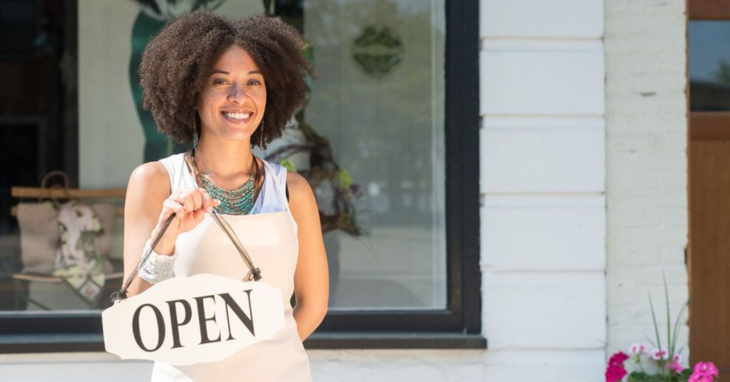 A woman in an apron stands holding white sign with black text reading "open"