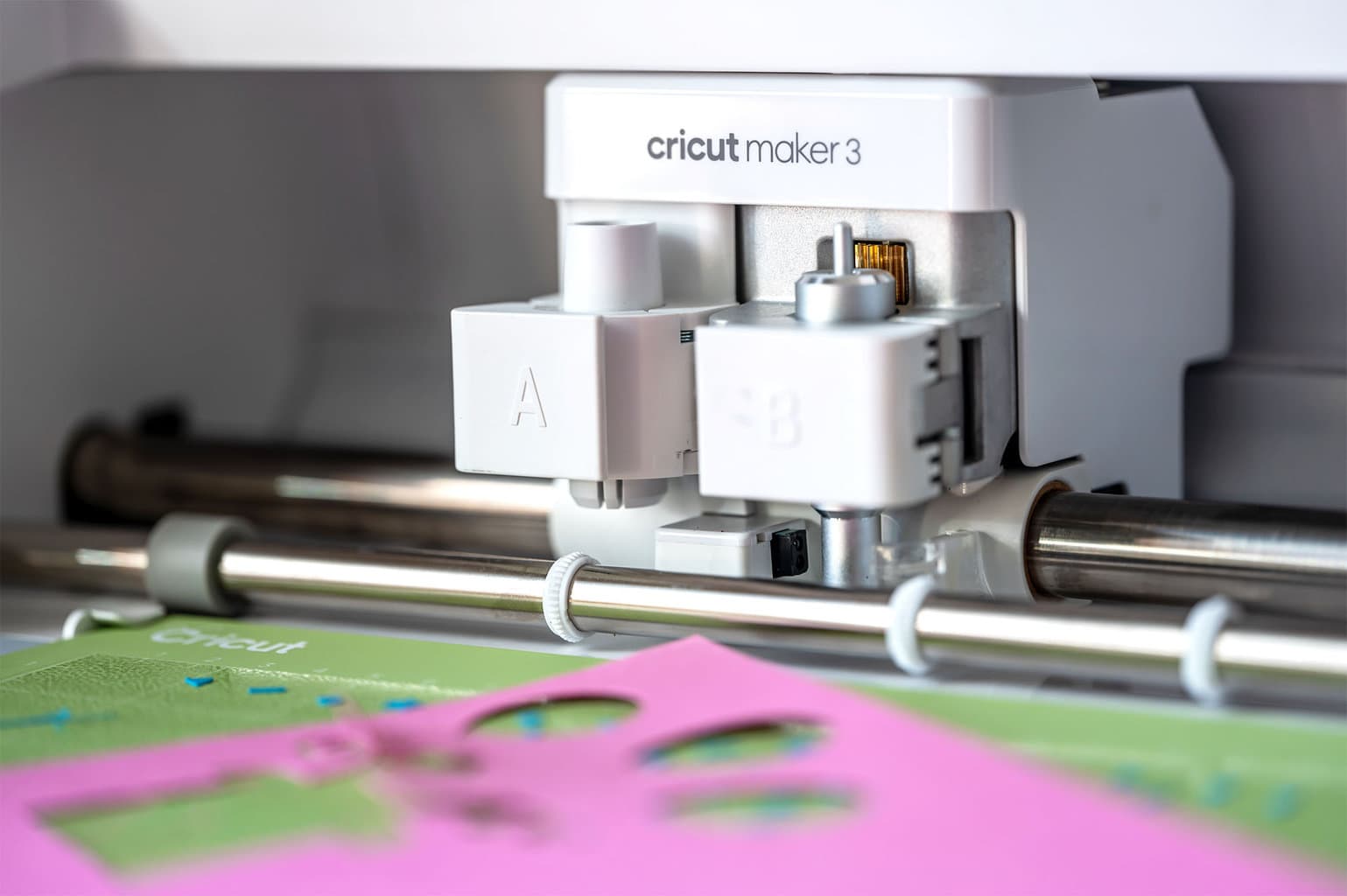 white cricut maker 3 cuts shapes out of a pink piece of cardstock with a green mat underneath