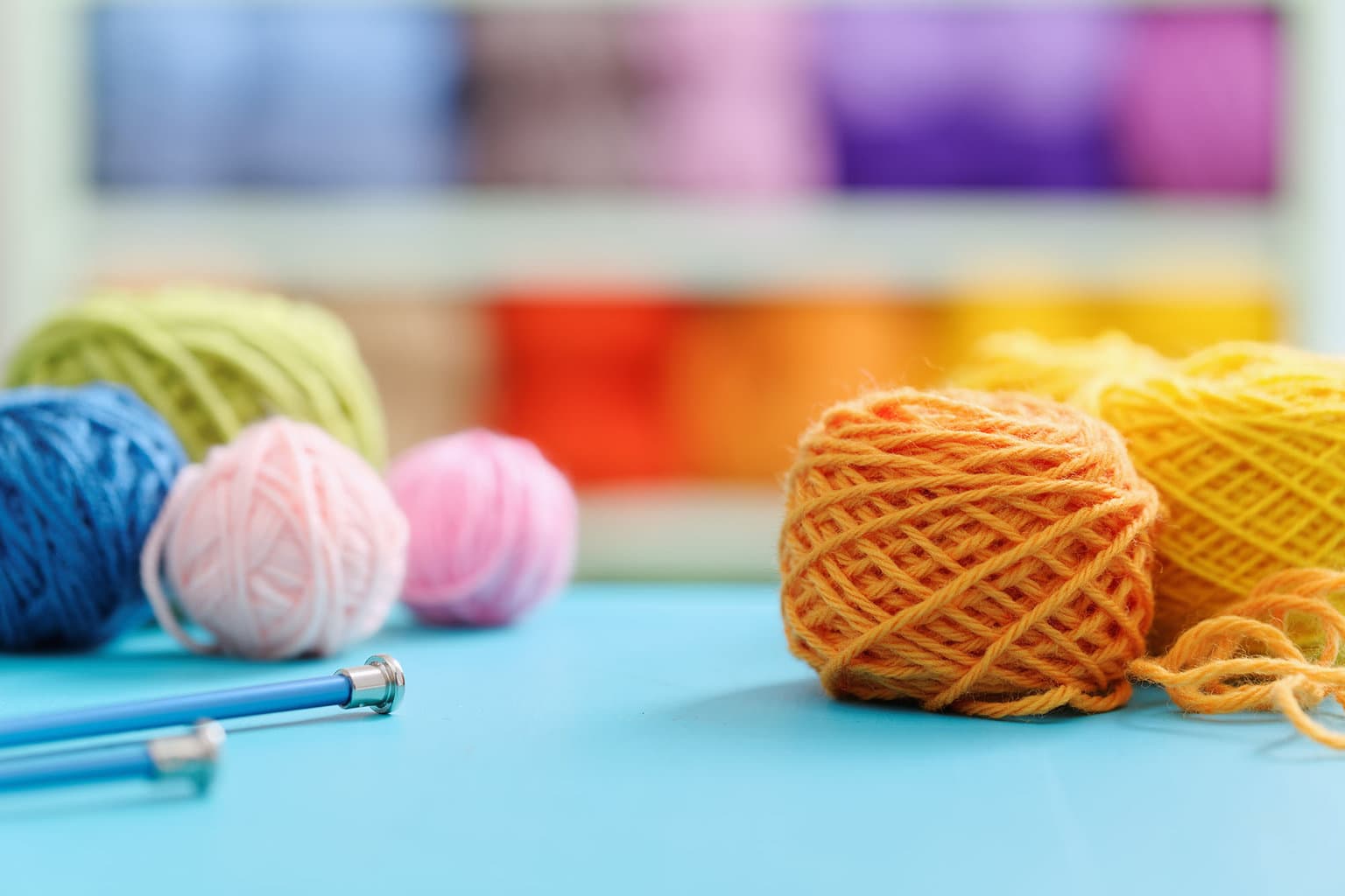 Several small balls of colorful yarn sit atop a robin's egg blue table with knitting needles to the side