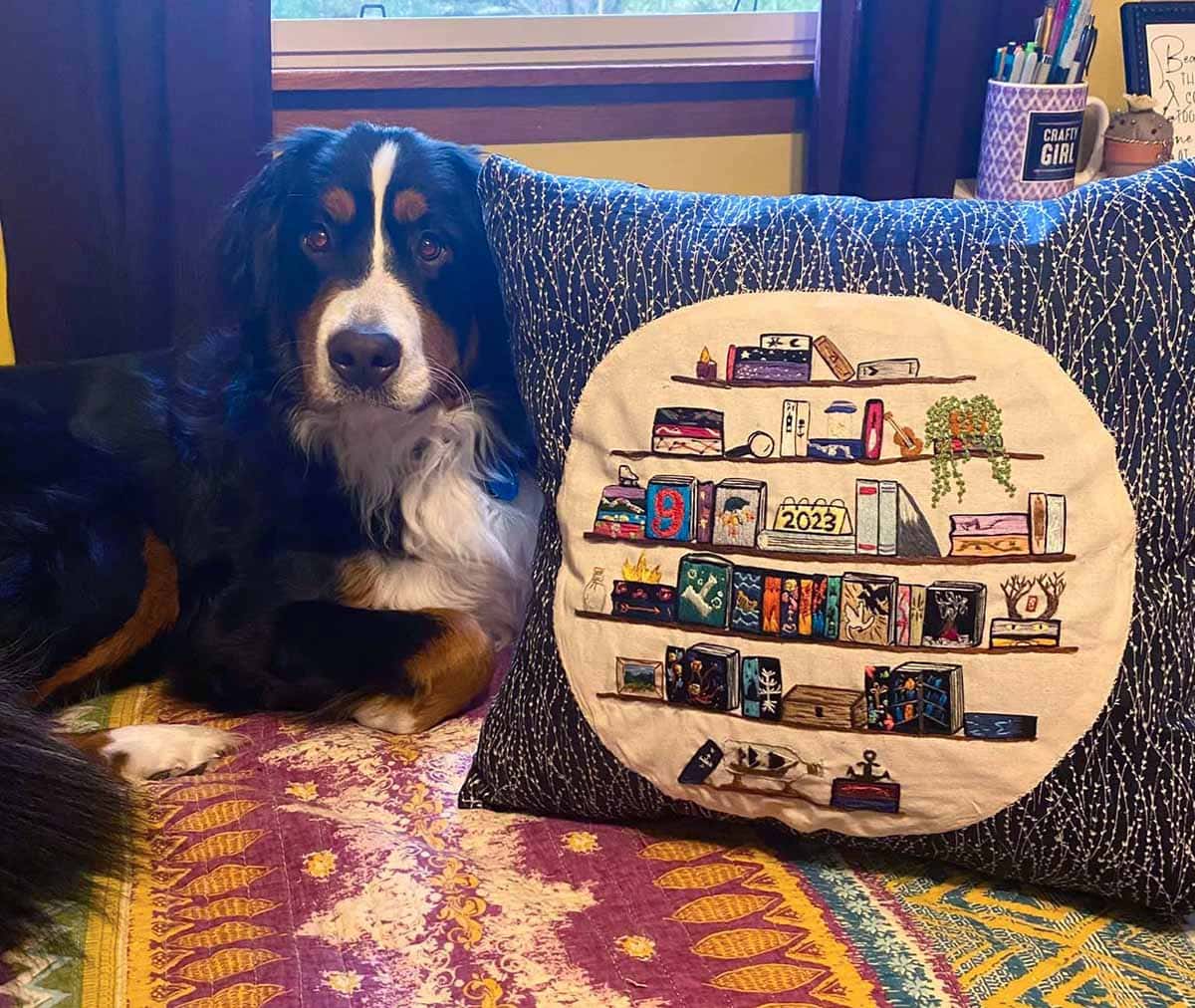 embroidered journal displays every book that the artist read for 1 year. it is featured on the front of a large blue pillow, which sits next to her Australian Shepard.
