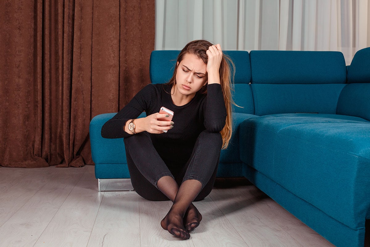 a woman dressed in all black sits in her living room floor looking at her phone with an expression of concern