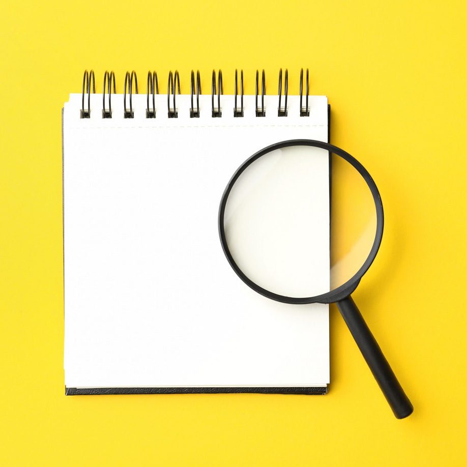 A large, black rimmed maginfying glass rests against a white, top bound spiral notebook. The background is a bright yellow.