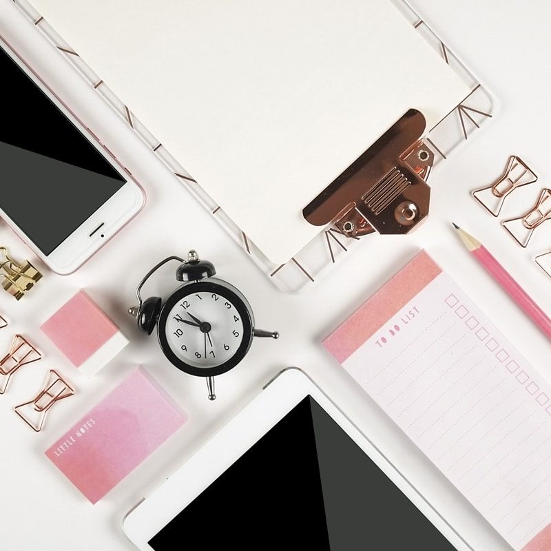 A diagonal arrangement of a clipboard, notepad, ipad, cellphone, binder clips, pencils and other office supplies with rose gold highlights.