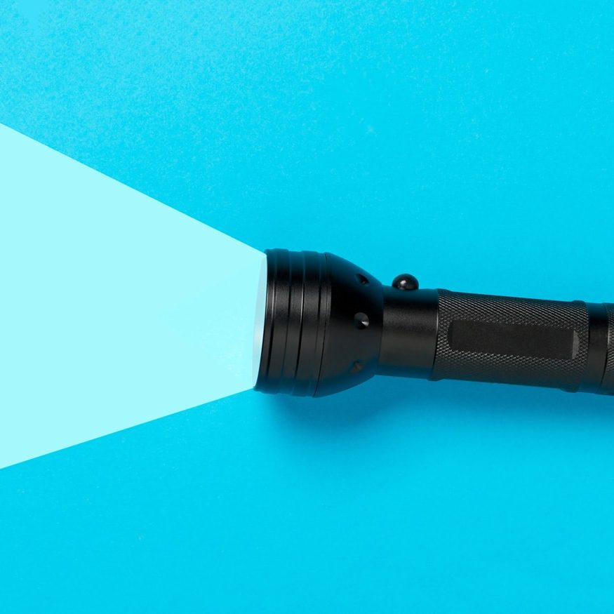 A flashlight is laying horizontal on the right side of the screen. The flashlight is black, and a light blue highlighted area is casting out of the flashlight as if it is lighting up the background. The other side of the background is a darker blue.